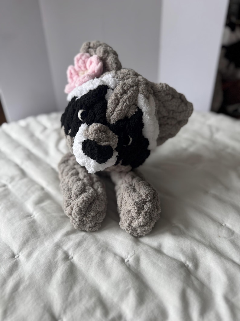 May include: A crocheted raccoon plush toy with a pink flower on its head. The toy is gray, black, and white and has a sweet expression.
