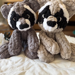 May include: Two plush crocheted raccoon toys with black eyes, black noses, and white markings on their faces. The toys are sitting on a white bedspread.