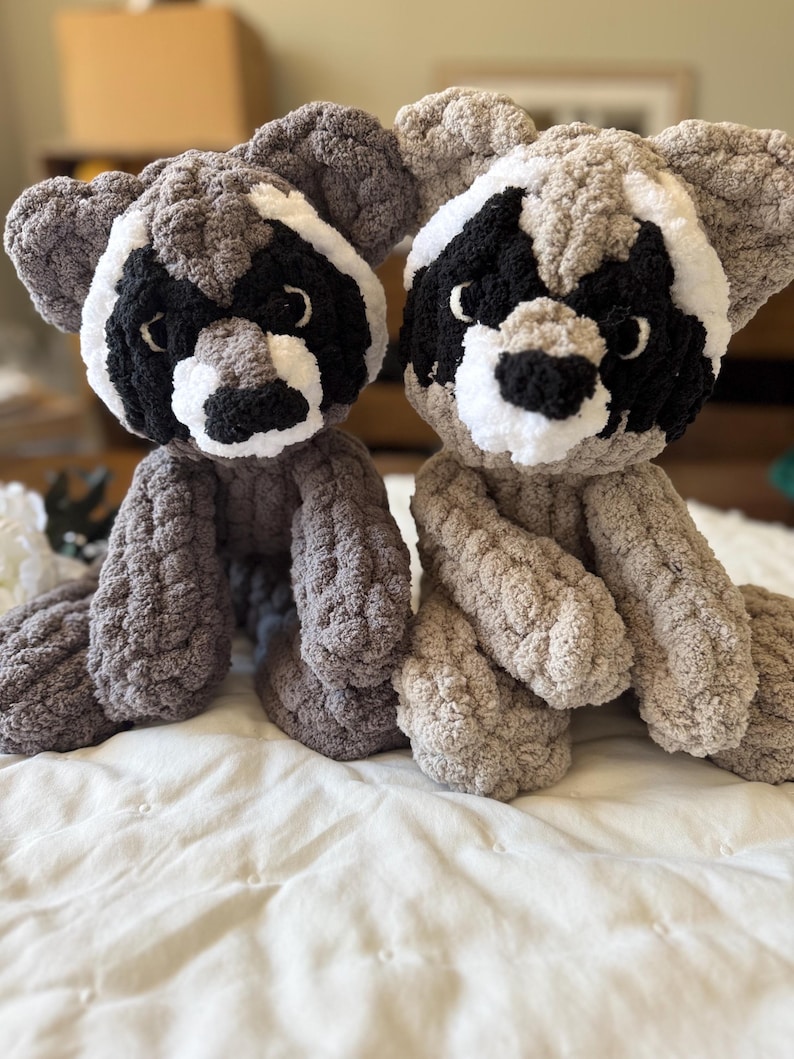 May include: Two plush crocheted raccoon toys with black eyes, black noses, and white markings on their faces. The toys are sitting on a white bedspread.