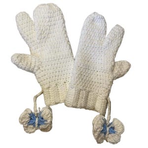 Op de afbeelding: Een paar witte gehaakte wanten met een ontwerp met drie vingers. Elke want heeft een klein, lichtblauw en wit vlinderaccent bevestigd aan de pols. De wanten zijn gemaakt van een getextureerd, gebroken wit garen.