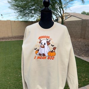 May include: A beige crewneck sweatshirt featuring an embroidered graphic of a brown highland cow dressed as a ghost. The design includes orange pumpkins, small black bats, and the text MOOOOO... I MEAN BOO in orange lettering.