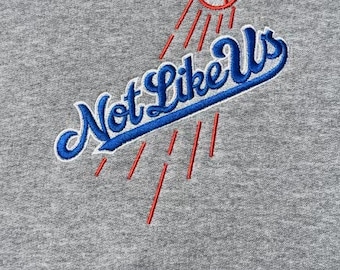 May include: Gray sweatshirt with a blue and red embroidered baseball design. The design features a baseball with red stitching, red rays of light, and the text "Not Like Us" in blue with a white outline.