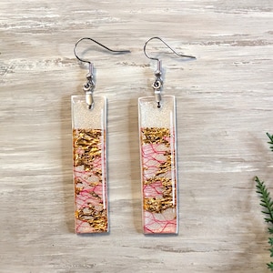 May include: A pair of rectangular dangle earrings. The top section is glittery silver, leading to gold leaf and pink thread details. The earrings are suspended from silver-coloured fish hook ear wires.