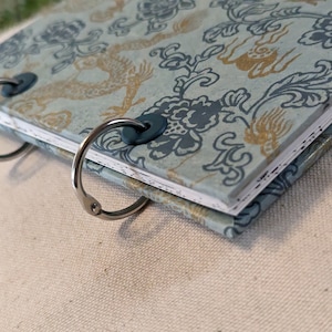 May include: A small, open notebook with a light blue cover featuring a floral and dragon pattern in gold and dark blue. The pages are held together by two silver binder rings. The notebook is on a light-colored surface.