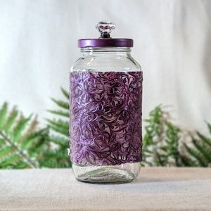 May include: A clear glass jar with a decorative purple band and lid. The band features a swirling pattern. The lid is a matching purple with a crystal-like knob. The jar is on a light-colored surface.