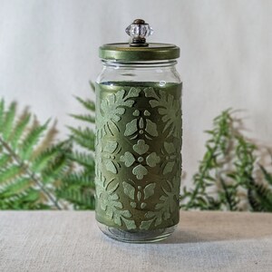 May include: A clear glass jar with a decorative olive green floral design. The jar has a matching lid and a clear crystal knob. The jar is a decorative storage container.