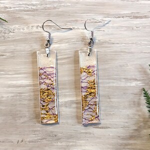 May include: A pair of rectangular dangle earrings. Each earring has a clear resin rectangle with gold and purple accents. The earrings have silver-coloured fish hook ear wires.