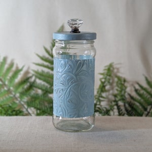 Könnte beinhalten: Ein klares Glas mit hellblauem Deckel und einem dekorativen hellblauen Band mit Wirbelmuster. Der Deckel ist mit einem klaren, facettierten Knauf versehen. Das Glas wird vor grünem Farn präsentiert.