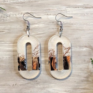 May include: A pair of oval drop earrings. The earrings have a silver glittery background with a central cutout. The cutout reveals a design with brown, black, and orange abstract shapes and lines. The earrings have silver hooks.