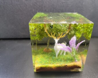 May include: A clear cube filled with resin contains a miniature forest scene. A white deer figurine stands in the centre, surrounded by green moss and purple flowers.