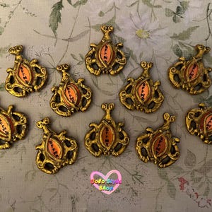 May include: A collection of ten ornate, gold-colored pendants. Each pendant features an orange center with black text and a detailed, swirling design. The pendants are arranged on a floral patterned surface.