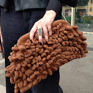 May include: A brown, looped-design clutch bag. The bag is held by a person wearing a black jacket and dark jeans. The clutch has a soft, plush appearance and a unique, eye-catching texture. The bag's texture is made of loops.