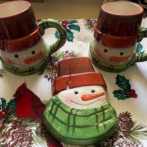May include: Three ceramic snowman mugs with green and brown plaid designs. The mugs are on a white tablecloth with a green and red holiday pattern.