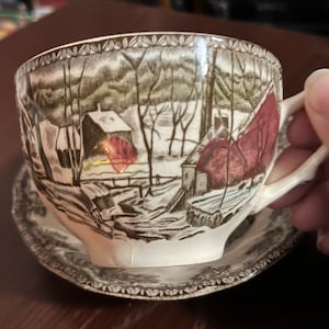 May include: A white teacup and saucer with a brown and red winter scene design. The scene features a small house with a red roof and a snowy landscape.