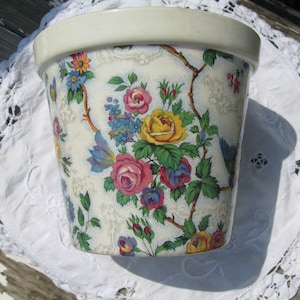 May include: A white ceramic flower pot with a floral pattern featuring pink, yellow, blue, and green flowers. The pot has a white background with a delicate lace-like design.