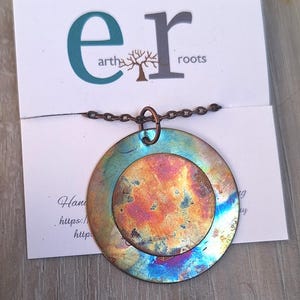 May include: A handcrafted pendant necklace featuring a large, iridescent, circular pendant with a smaller, central circle. The pendant hangs from a delicate chain. The card behind the pendant has the text "earth roots".