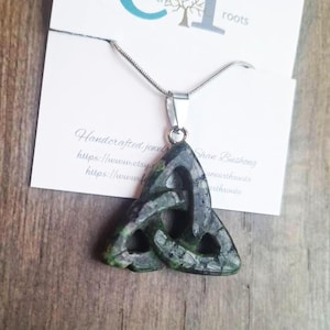 May include: A handcrafted green and gray Celtic knot pendant necklace. The pendant is made of stone and hangs from a silver chain. The necklace is displayed on a white card with the text "arth roots".