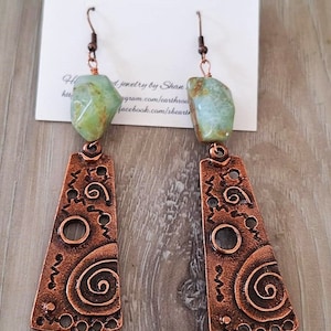 May include: Copper earrings with a green stone dangle. The earrings feature a copper pendant with a swirl design and a green stone.