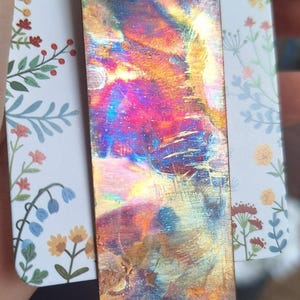 May include: A rectangular pendant featuring a heat-treated metal surface with a vibrant, iridescent finish displaying shades of blue, purple, magenta, and gold. The pendant hangs from a grey cord and is attached to a white card with floral patterns.