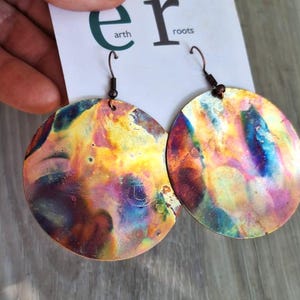 May include: A pair of large, round earrings with a colorful, iridescent finish. The earrings display hues of yellow, orange, blue, and purple. The ear wires are dark brown. The earrings are hanging in front of a white card with the words "earth roots".