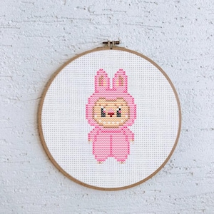 Op de afbeelding: Een roze kruissteek borduurring met een cartoon konijn karakter in een roze jumpsuit met een capuchon en oren. Het karakter heeft een klein gezicht met zwarte ogen en een rode neus. De borduursel is tegen een witte achtergrond.