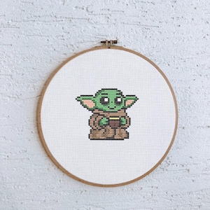Peut inclure: Un cerceau de broderie en point de croix avec une image pixelisée en vert et marron d'un personnage de la franchise Star Wars, connu sous le nom de Baby Yoda, tenant une tasse.