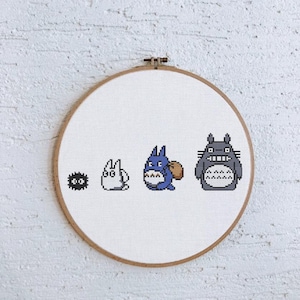 May include: A cross-stitch embroidery hoop featuring four characters from the animated film "My Neighbor Totoro." The characters are stitched in black, white, blue, and gray on a white fabric background. The wooden hoop is visible.
