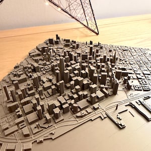 May include: A 3D-printed model of a city, featuring detailed buildings and streets. The model is in shades of gray and brown, with a textured surface. A decorative star-shaped light fixture hangs above the model, adding a warm glow.