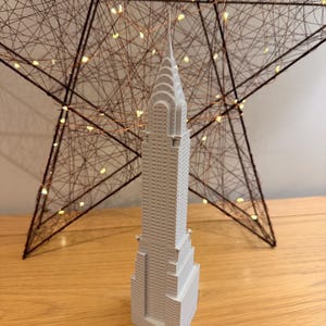 May include: A gray miniature replica of the Chrysler Building, standing on a wooden surface. Behind it is a large, star-shaped decoration made of intertwined brown wires, illuminated by small, warm-toned lights. The building has detailed architectural features.