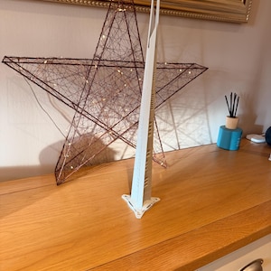 May include: A tall, off-white, three-sided tower sculpture with a pointed spire and a triangular base. The structure features a series of horizontal cutouts along its vertical edges, creating a slender, architectural form that stands upright.