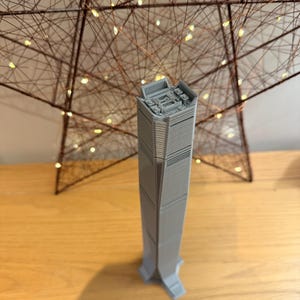 May include: A grey, 3D-printed model of a skyscraper with a detailed top section. The building is set against a backdrop of a brown star-shaped decoration with small, warm white lights. The model stands on a wooden surface.