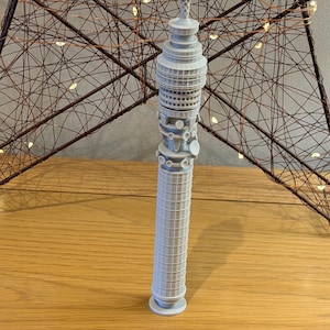 May include: A light gray miniature of the BT Tower, a prominent London landmark. The model features detailed architectural elements, including the tower's cylindrical base and the various antennas and structures at the top. The model is displayed on a wooden surface.