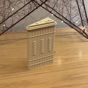 Peut inclure: Une réplique beige miniature du Flatiron Building. Le modèle présente des détails architecturaux, notamment des fenêtres et des lignes verticales, sur un fond neutre. Le bâtiment mesure environ 10 cm de haut.