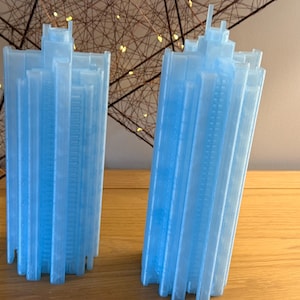 May include: Two light blue, translucent, 3D-printed skyscraper models featuring a multi-tiered architectural design with vertical columns and a central tower structure. Each model has a smooth, semi-glossy finish and a rectangular base footprint.