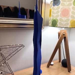 Può includere: Un modello di grattacielo stampato in 3D di colore blu. Il modello è alto e sottile, con un design geometrico unico. È posizionato su una superficie di legno.