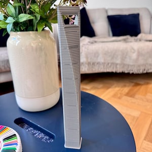 May include: A gray 3D printed model of a skyscraper, sitting on a blue table. The model is detailed and has a small window opening at the top.