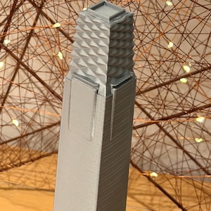 May include: A tall, grey, 3D-printed architectural model of a skyscraper. The structure features a smooth, rectangular base that transitions into a textured, geometric upper section with a repeating diamond pattern, topped by a flat square roof.