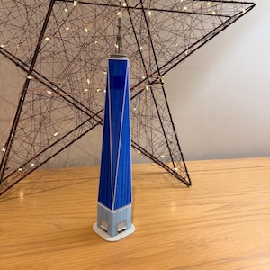 Könnte beinhalten: Ein detailliertes Miniaturmodell des One World Trade Centers, überwiegend blau mit weißen Details und einer silbernen Spitze. Das Modell steht auf einem weißen Sockel. Im Hintergrund befindet sich eine große sternförmige Dekoration mit Lichterketten.