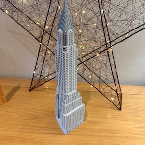 May include: A gray miniature model of the Chrysler Building, a famous skyscraper in New York City. The model is detailed with windows and architectural features. It stands on a wooden surface, with a star-shaped decoration in the background.
