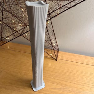 May include: A slender, pale grey vase with vertical ribbed detailing. The vase has a square base and a flared top. The vase is displayed on a wooden surface, with a decorative star-shaped object in the background.