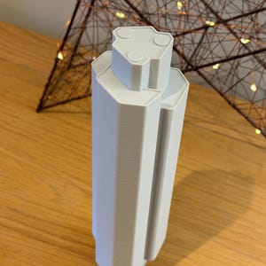 May include: A light gray, 3D-printed architectural model with a geometric design. The model has a rectangular base and a multi-faceted top with circular details. The object is displayed on a wooden surface.