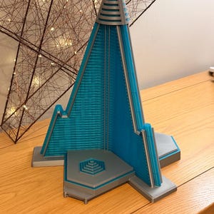 Può includere: Una scultura geometrica turchese e argento con una parte superiore triangolare e una base a gradini. La scultura ha un design architettonico moderno, con una facciata turchese strutturata e dettagli argentati. Si trova su una superficie di legno.