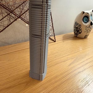 May include: A gray, miniature skyscraper model with a detailed facade. The building is on a wooden surface, with a decorative star-shaped object and a ceramic owl figurine in the background. The model is a detailed replica of a modern building.