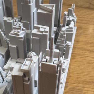 Peut inclure: Maquette grise imprimée en 3D d'une ligne d'horizon urbaine, avec de nombreux gratte-ciel et bâtiments de hauteurs et de conceptions variées. Le modèle est posé sur une surface en bois, mettant en valeur les détails architecturaux.