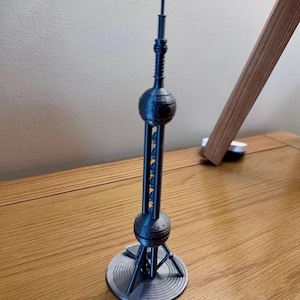 May include: A 3D-printed model of the Oriental Pearl Tower. The dark grey miniature features two spherical structures connected by a central column, supported by a circular base. The tower's antenna extends upwards, showcasing architectural detail.