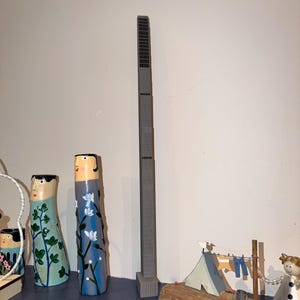 May include: A tall, slender, gray rectangular skyscraper model standing vertically. Beside it are two cylindrical vases with painted faces and floral patterns, and a small wooden base featuring a miniature tent and laundry line display.