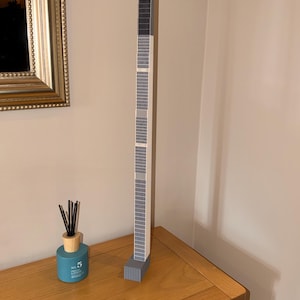 May include: A tall, slender, vertical model of a skyscraper constructed from stacked rectangular blocks. The structure features a repeating grid pattern of dark grey and light grey segments, culminating in a solid dark grey top section and a base.