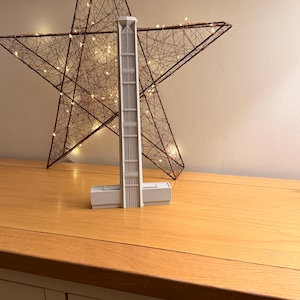 May include: A light grey architectural model of a building with a tall, rectangular tower and a horizontal base. The model is displayed on a wooden surface, with a decorative star-shaped light fixture in the background.