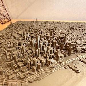 May include: A detailed, three-dimensional model of a city, showcasing a dense urban landscape with numerous buildings and streets. The model is a light brown color, with a textured surface, and appears to be made of a solid material.