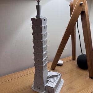 Puede incluir: Un modelo de rascacielos impreso en 3D, pintado en color gris claro. El modelo está detallado con múltiples niveles y una parte superior puntiaguda.